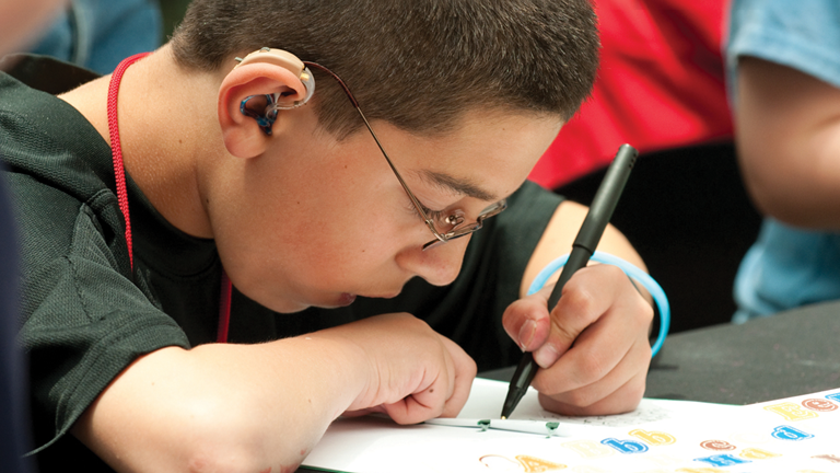 A boy with short brown hair wearing a hearing aid and glasses with a light blue wrist band and black t-shirt is drawing on a piece of paper with a pen he is holding in his left hand.