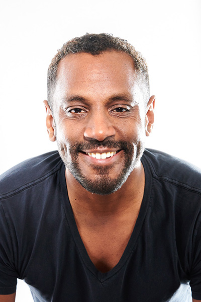 A headshot of composer Carlos Simon. Smiling, he looks towards the camera while wearing a black t-shirt in front of a white background.