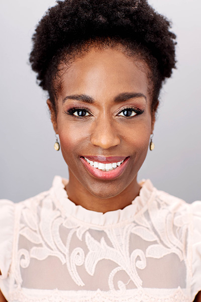 A headshot of artist Felicia Curry. Smiling, she wears a light lacey top with earrings in front of a gray background.