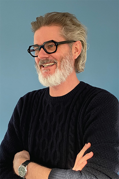 A headshot of Mo Willems. Smiling with his arms crossed, he wears a black sweater, black eyeglasses, and a black watch while standing in front of a blue background.