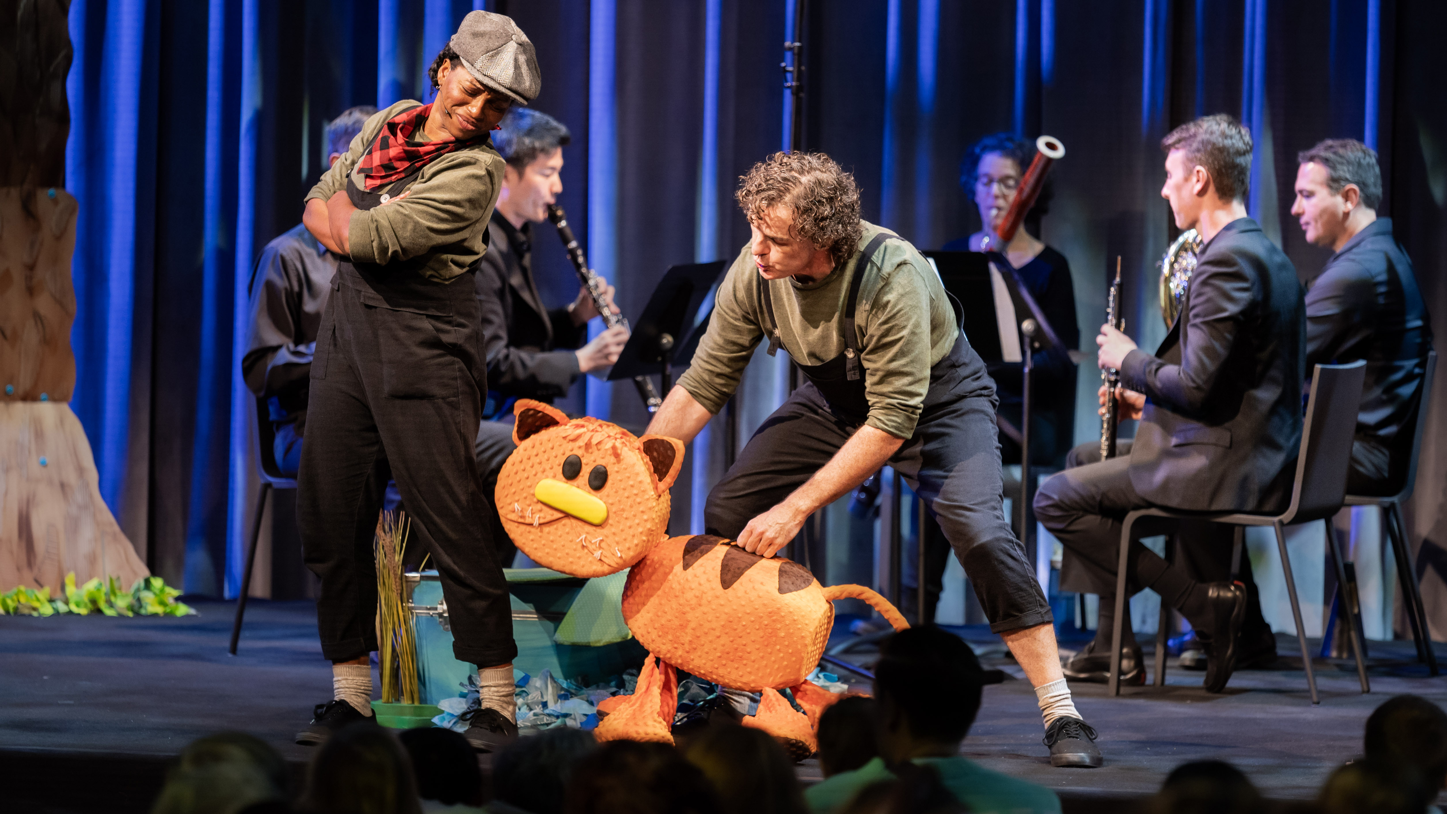 Two actors in dark green shirts and black overalls perform in front of an audience of children and adults. The standing performer wears a black hat and red-and-black plaid neck scarf, arms crossed while looking down at Cat. The crouching performer controls the Cat puppet, which is orange with brown stripes and a yellow nose. Five musicians in all-black outfits sit on chairs in a semi-circle behind the performers while playing instruments.