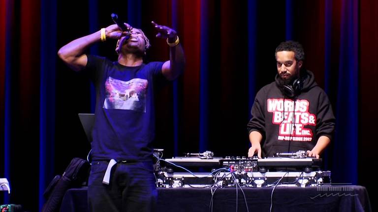 Javier Starks on stage with a microphone in hand performing at the Kennedy Center in 2015 with a DJ at the turntables to Javier’s left who has the words and logo for Words, Beats, and Life Incorporated on his sweatshirt.