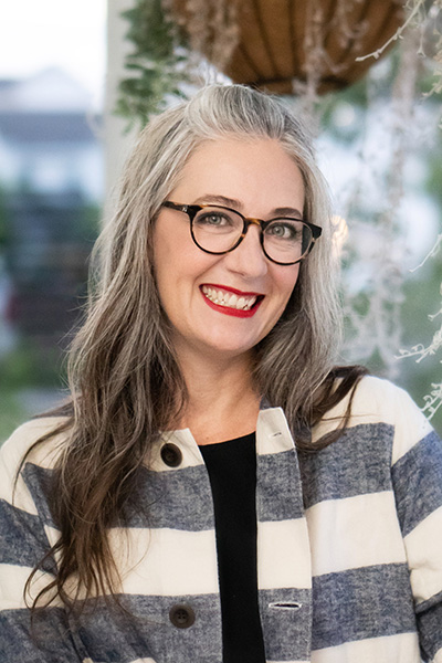 A headshot of dramaturg Megan Alrutz taken in an outside location. Smiling, she wears a white-and-gray striped jacket over a black shirt and dark eyeglasses.