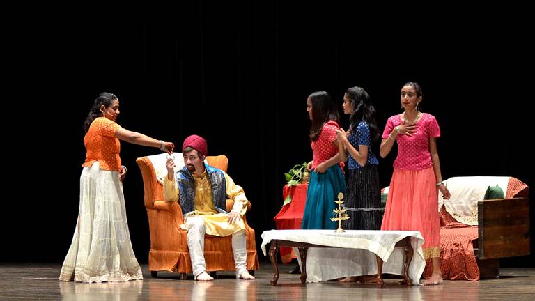 A stage set with actors in traditional Indian dress acting in Cinderella. To the left, a smiling woman is showing a bangle to a surprised man sitting in a chair. To the right, three other women look at them with concern.