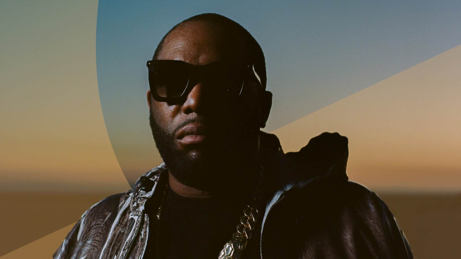 Killer Mike wearing sunglasses and a cool jacket in front of a muted sunset