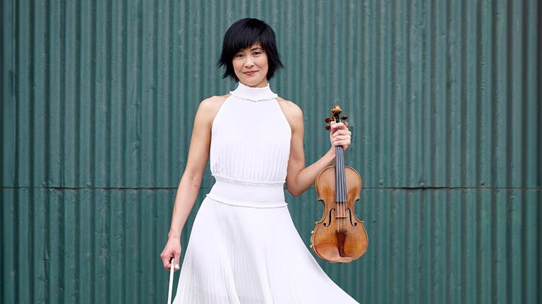 Jennifer Koh ins  white dress, holding her violin. She is standing in front of dark teal background.