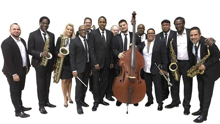 Jazz Houston Orchestra performers in front of a white background. There are all in black tie and facing the camera. Various performers are holding instruments including a double bass and brass intruments.